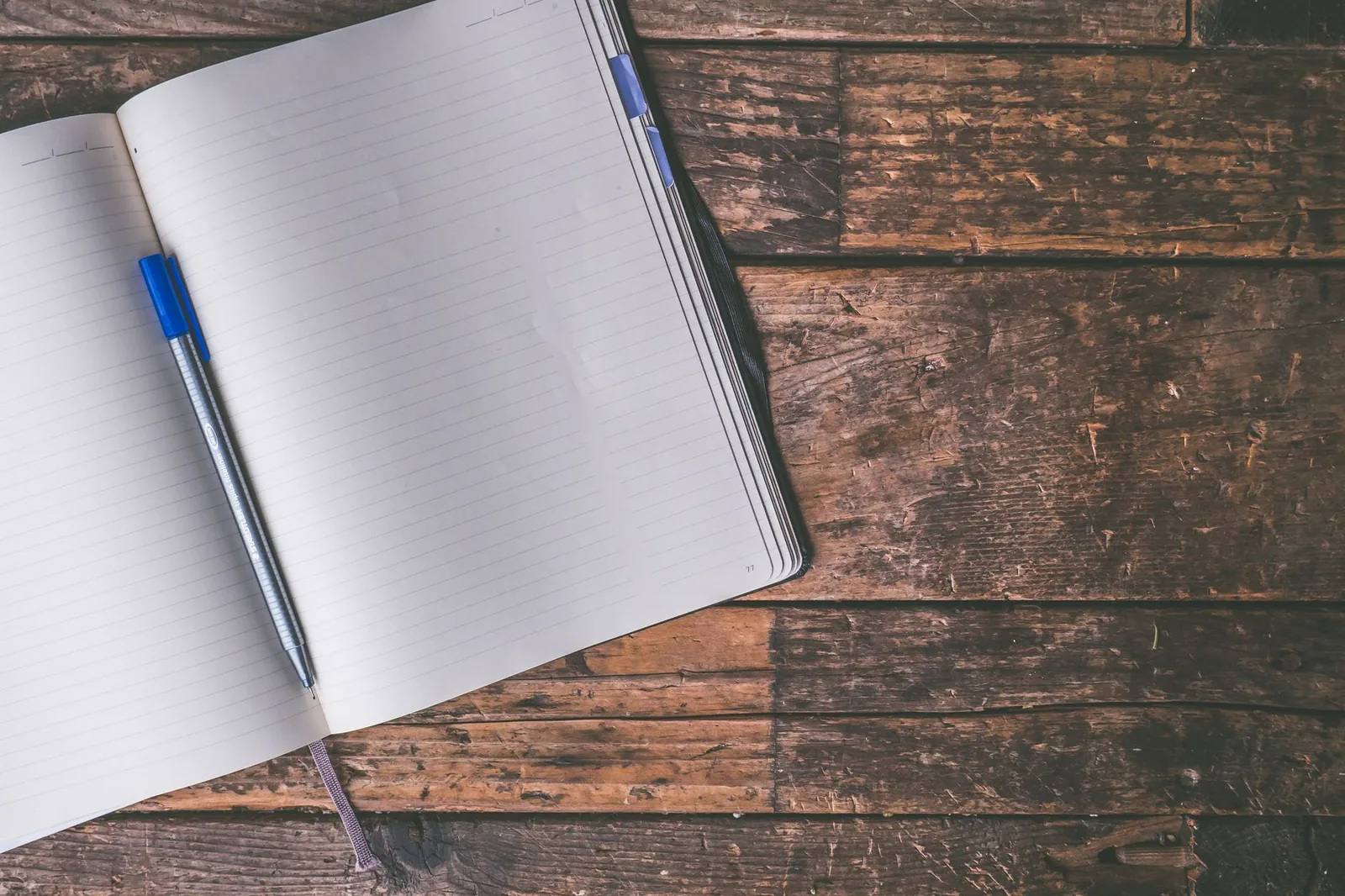 An open lined notebook and a blue ballpoint pen resting on a rustic wooden table.